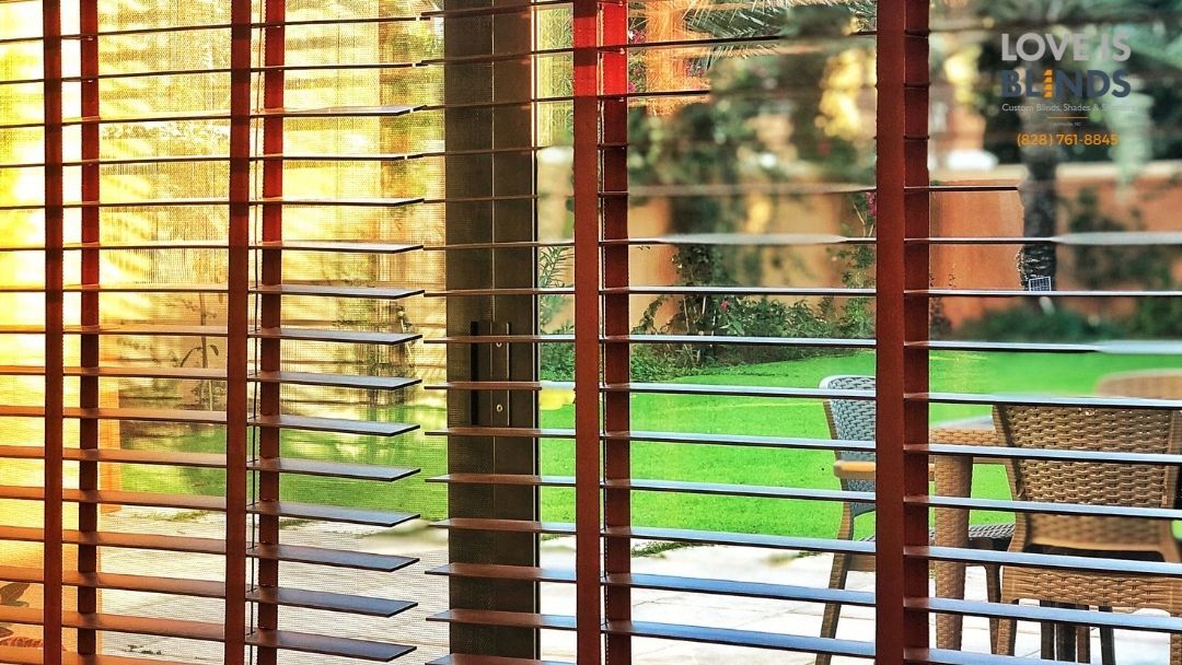 Sunlit patio seen through wooden blinds, with green lawn and a wicker chair outside.