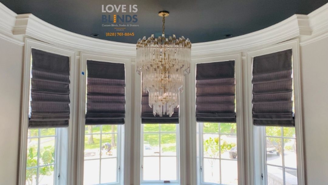 Elegant bay window with white trim, gray Roman shades, and a crystal chandelier in a bright room