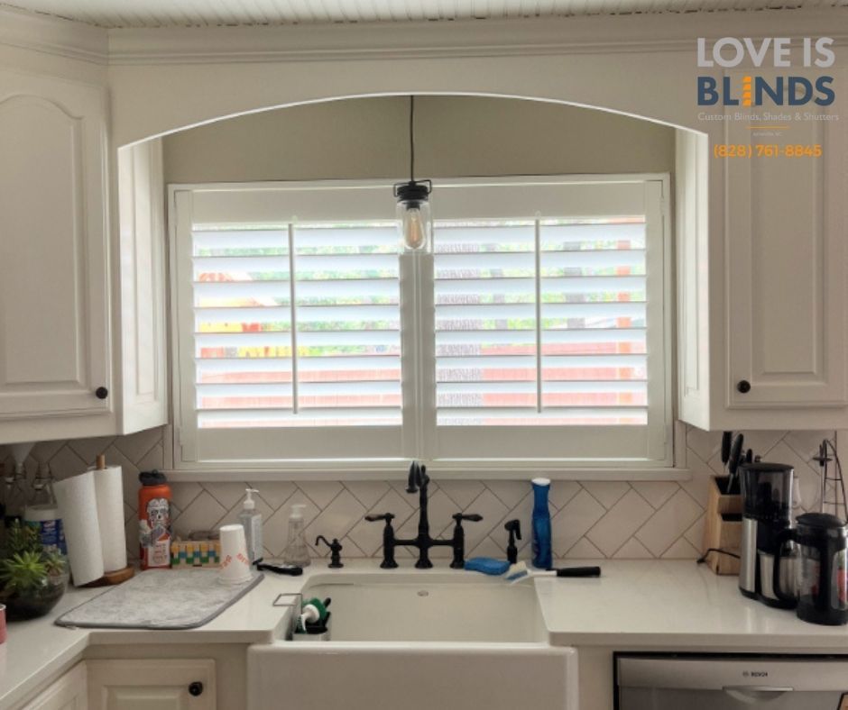 Bright kitchen sink under arched window with white shutters, black faucet, and white cabinets