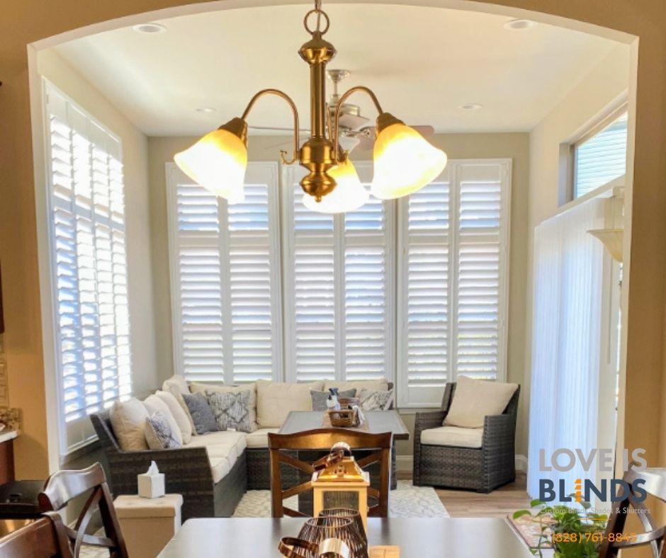 Bright sunroom with white shutters, gray seating, and a brass chandelier over a wooden table.