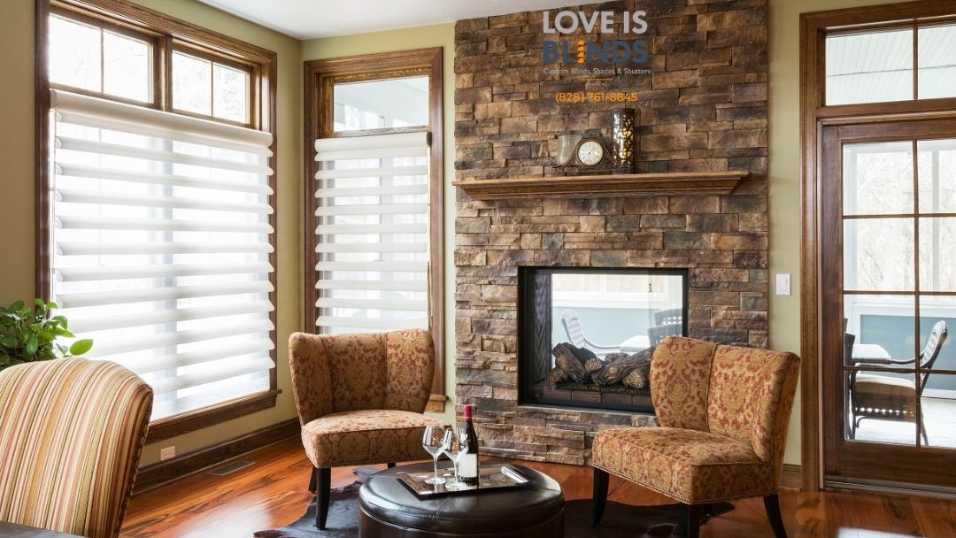 Cozy living room with stone fireplace, two patterned chairs, and large windows with blinds