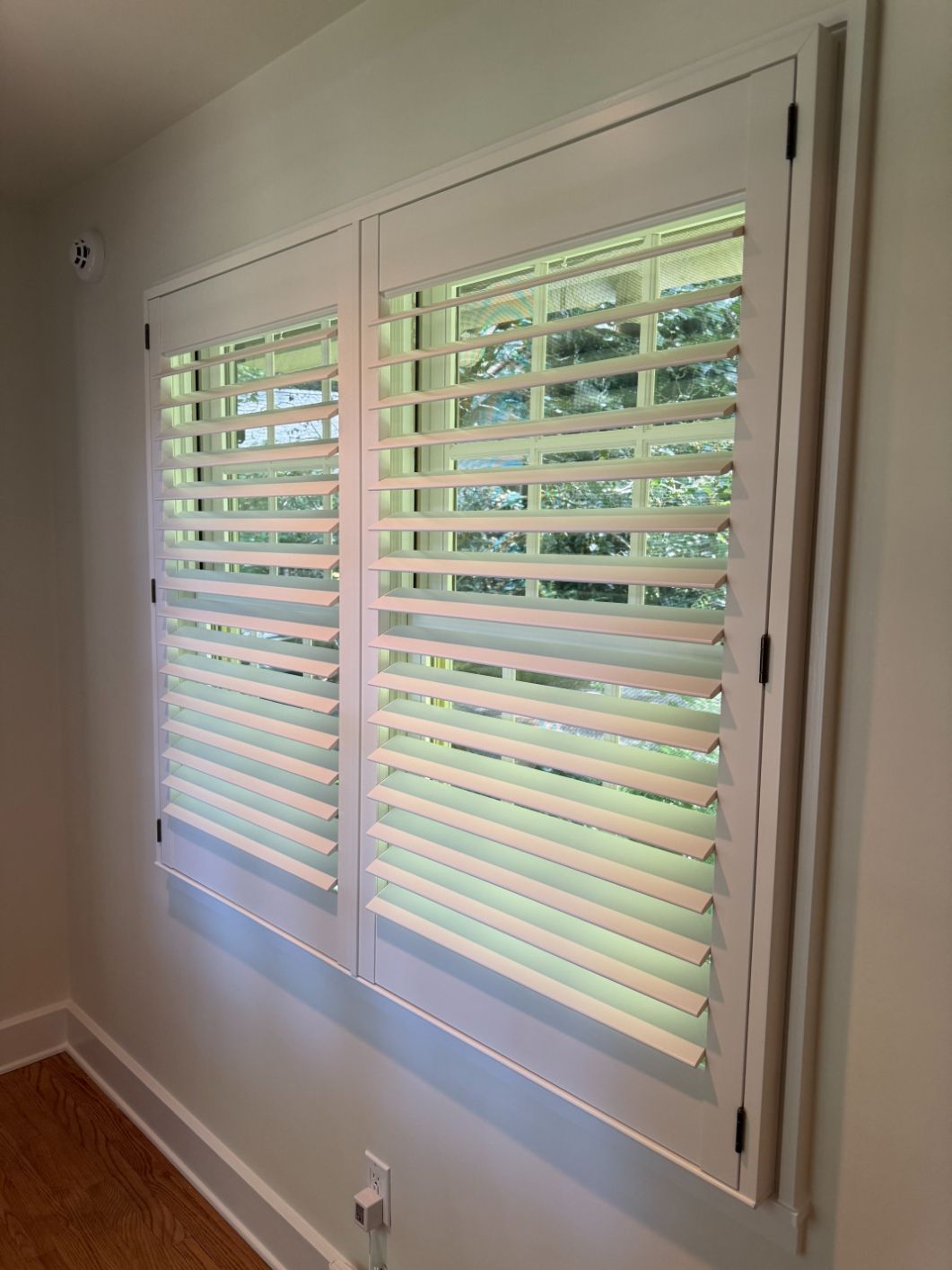 White window shutters with horizontal slats in a beige-walled room, showing greenery outside.