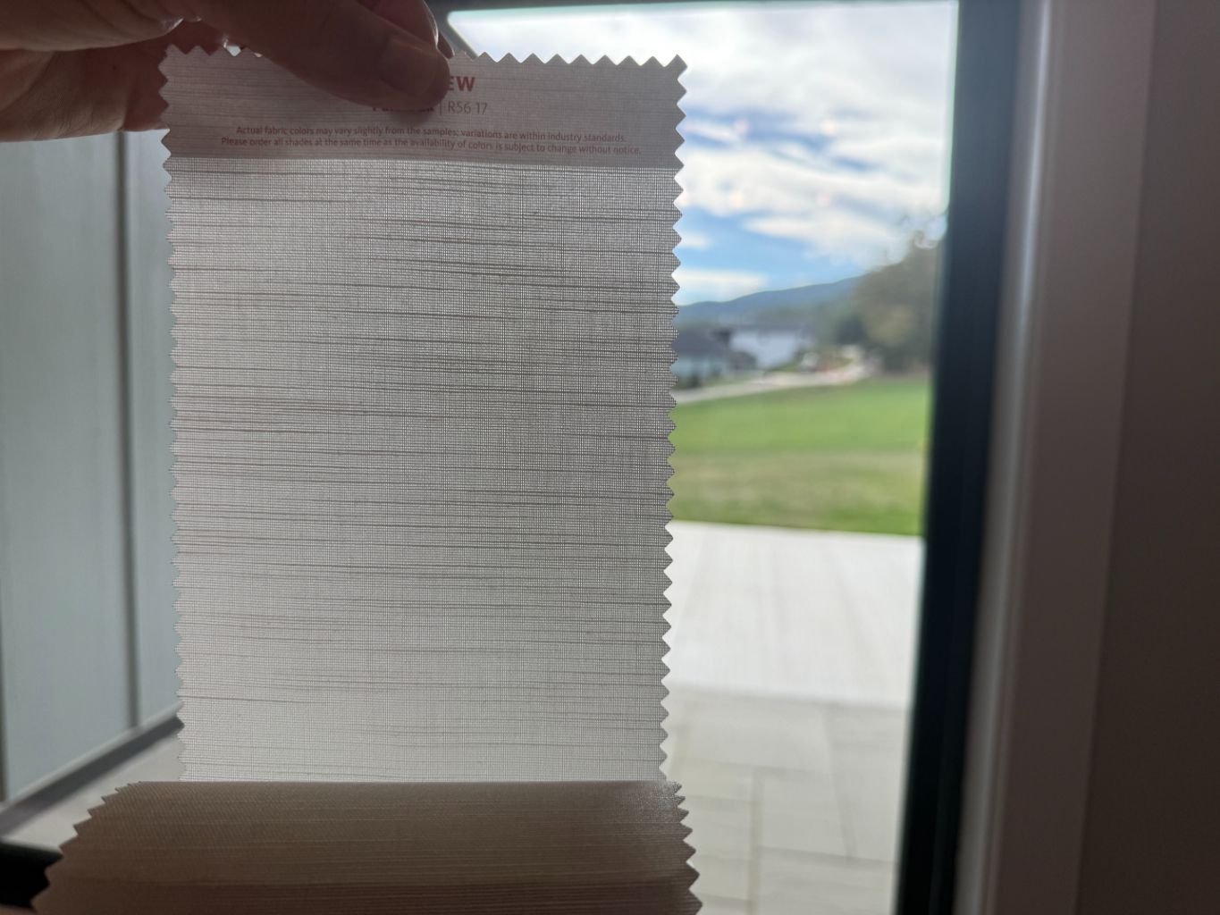 Hand holding a translucent woven window shade in front of a bright outdoor view