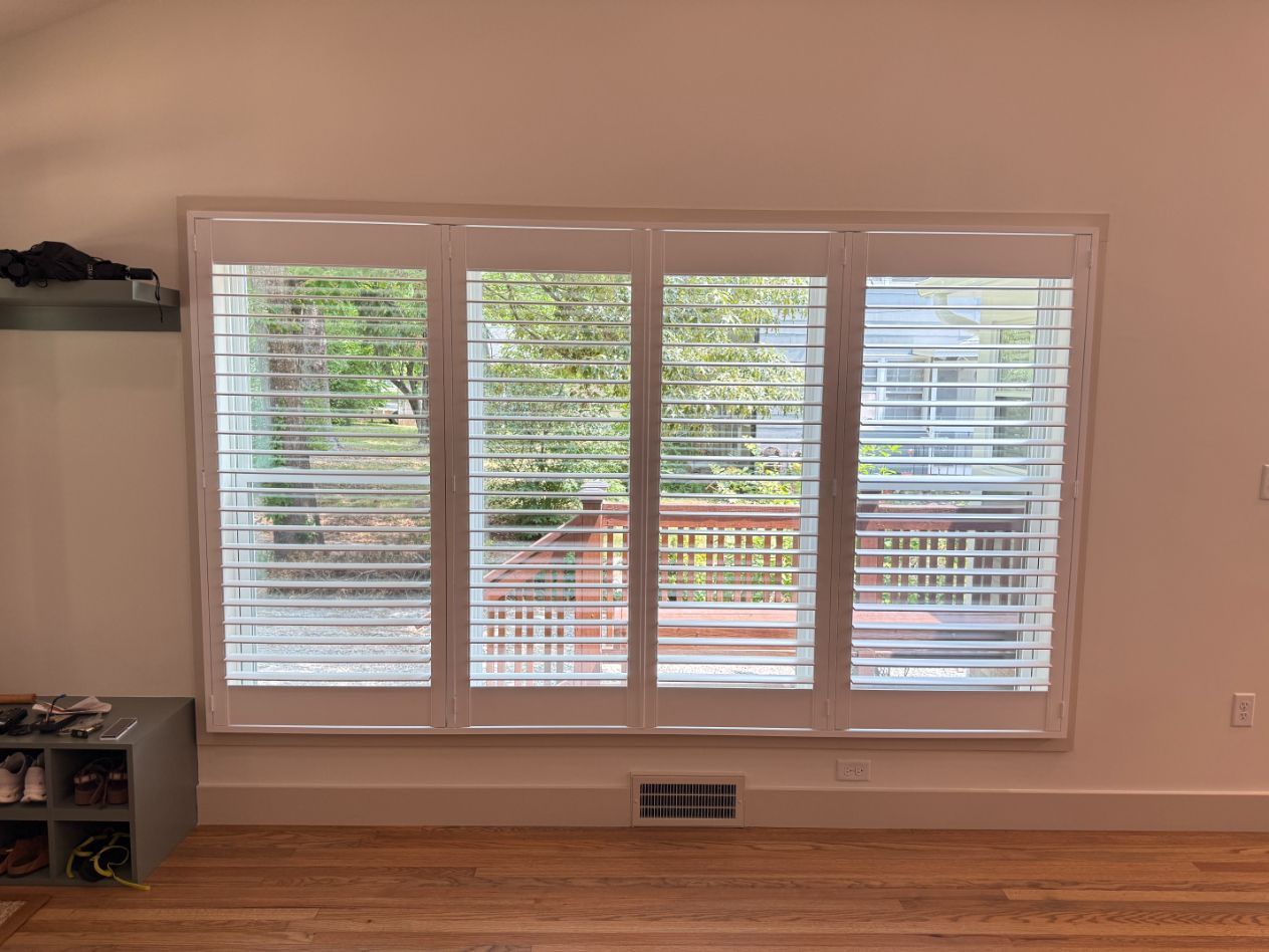 Empty living room with large white plantation shutters and a view of trees and deck outside