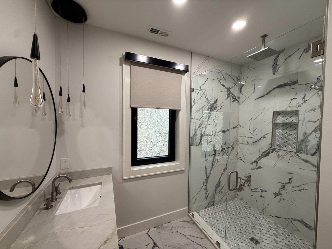Modern marble-tiled bathroom with glass shower, vanity, and window roller shade.