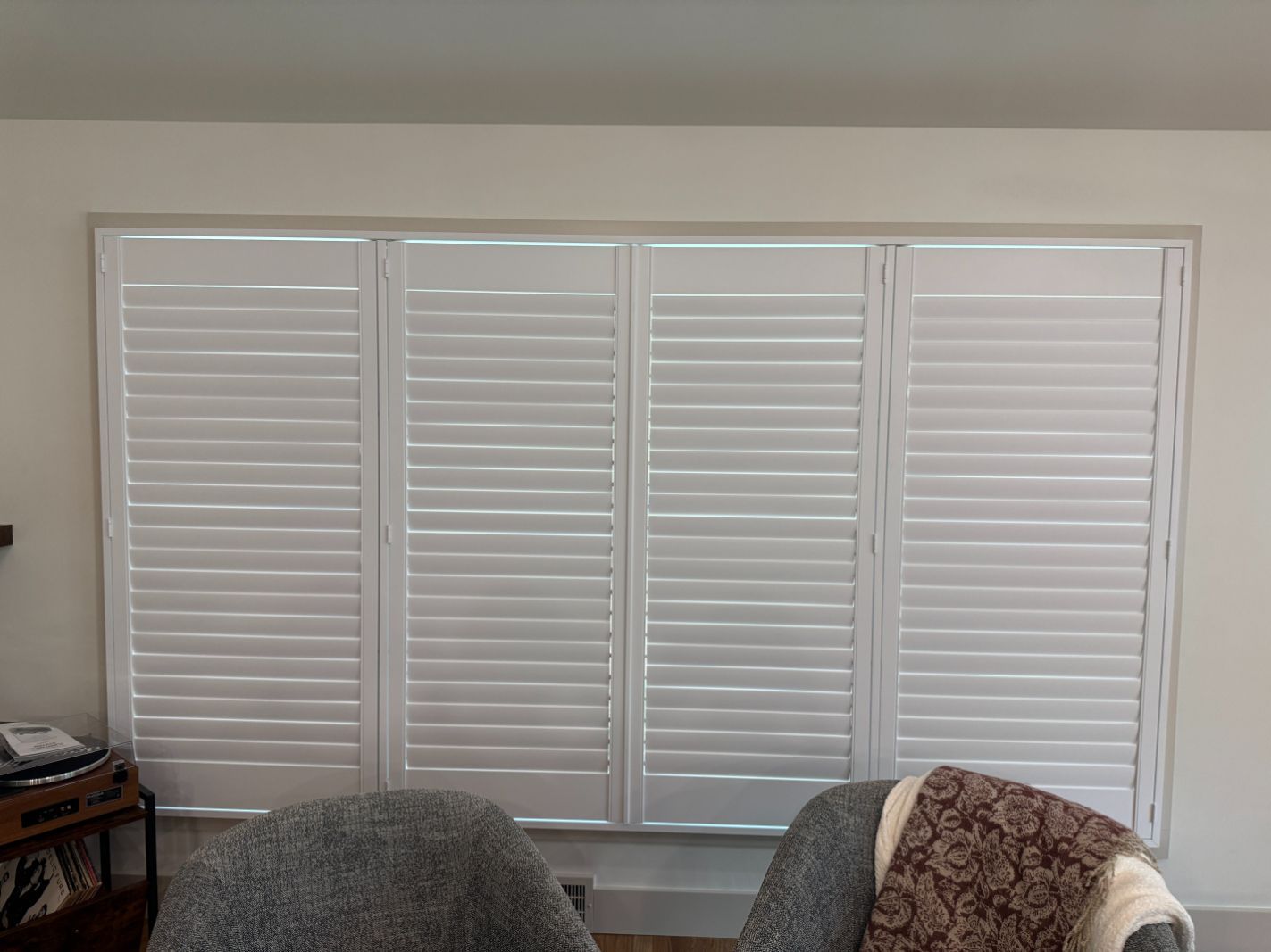 White plantation shutters on a large window in a bright room with two chairs below.