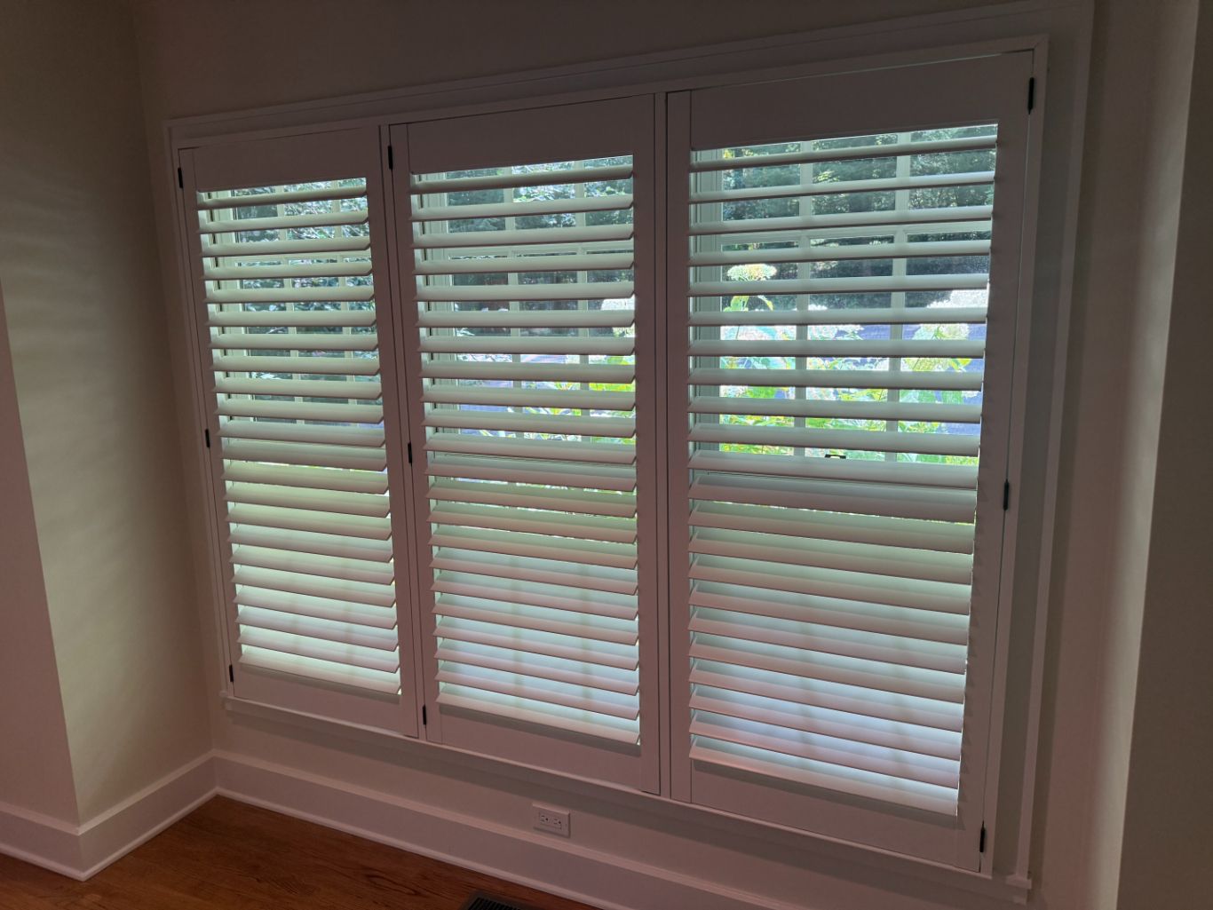 White plantation shutters on a large window in a beige room, with a view of greenery outside