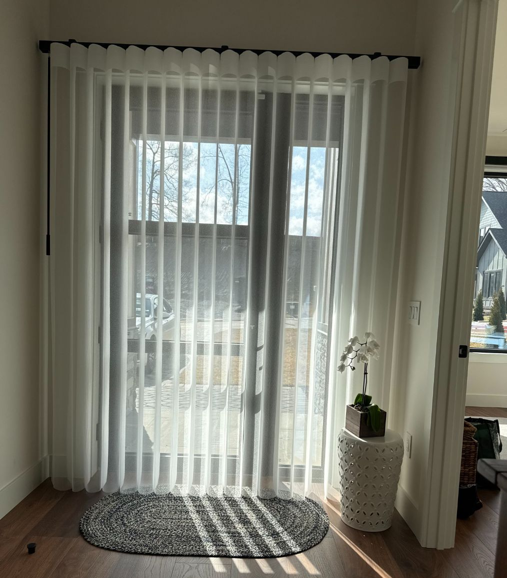 Sheer curtains cover a glass door, with a small rug and potted plant by a bright window.