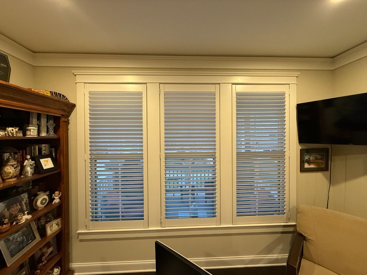 Living room with large windows, blinds, wall-mounted TV, and a wooden cabinet with dishes