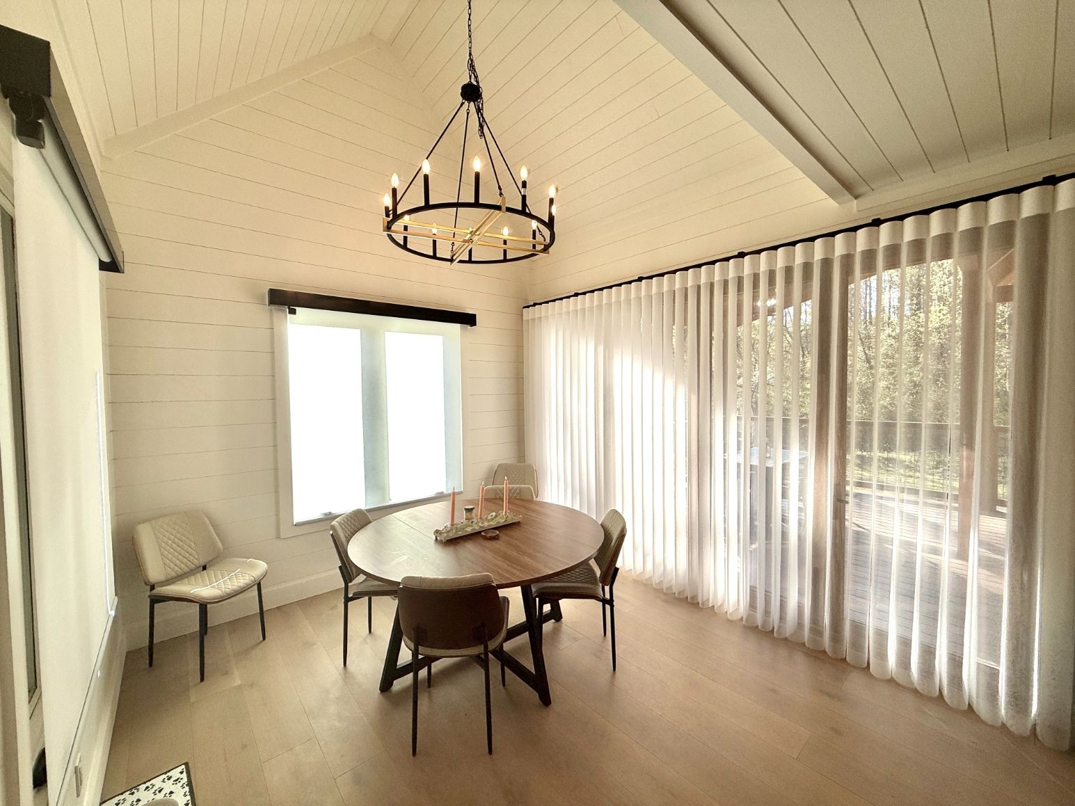 Bright sunroom with round dining table, chairs, chandelier, and sheer curtains by large windows