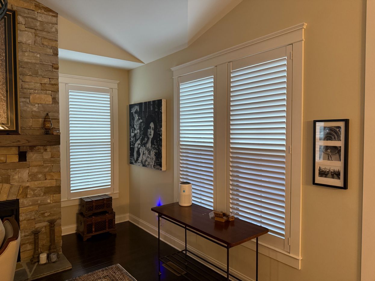 Living room corner with windows, white blinds, wall art, and a console table beside a stone fireplace.