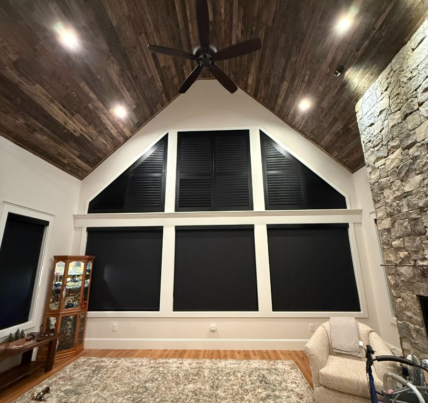 Vaulted living room with dark wood ceiling, ceiling fan, stone wall, and large black-shaded windows