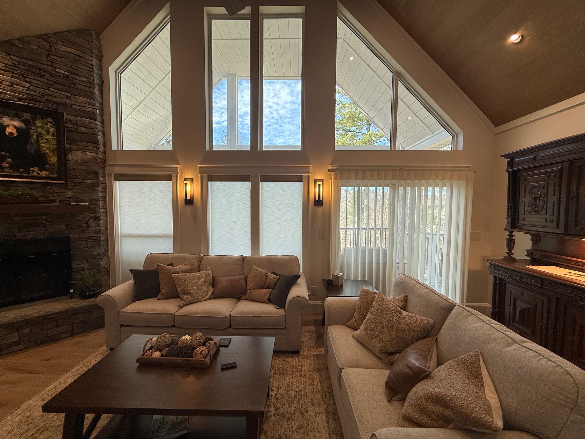 Spacious living room with vaulted ceiling, large windows, beige sofas, coffee table, and stone fireplace