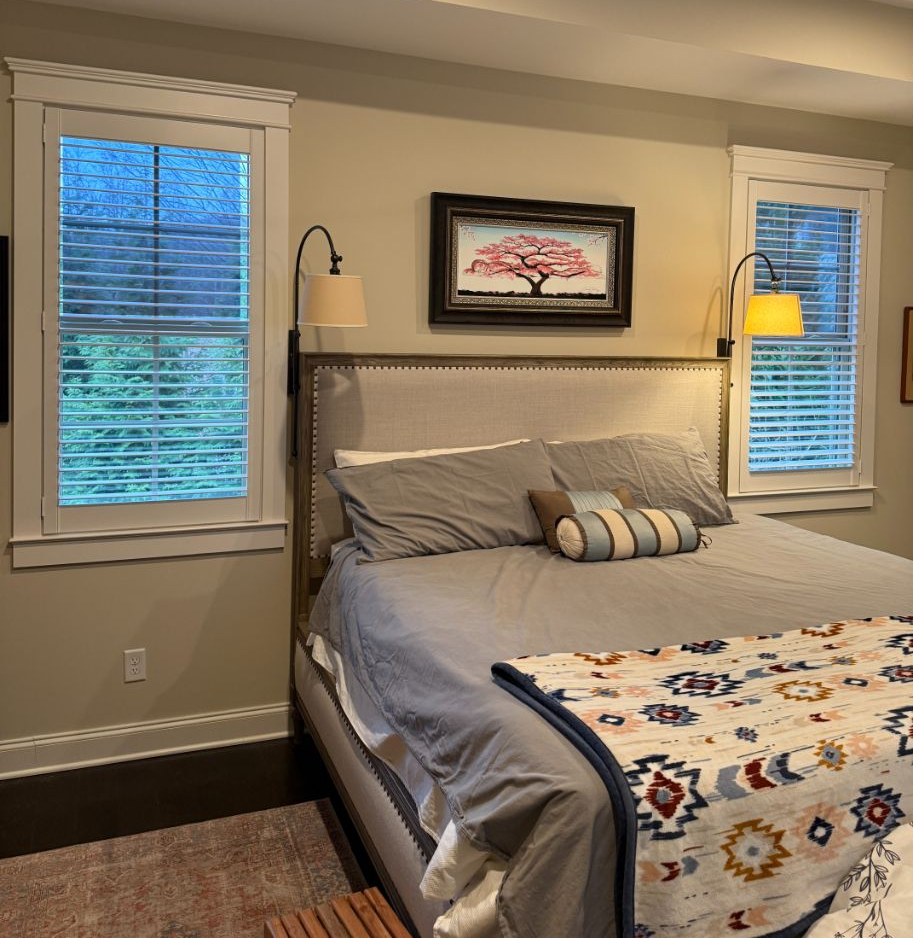 Bedroom with gray bed, patterned blanket, framed wall art, and two windows with blinds.