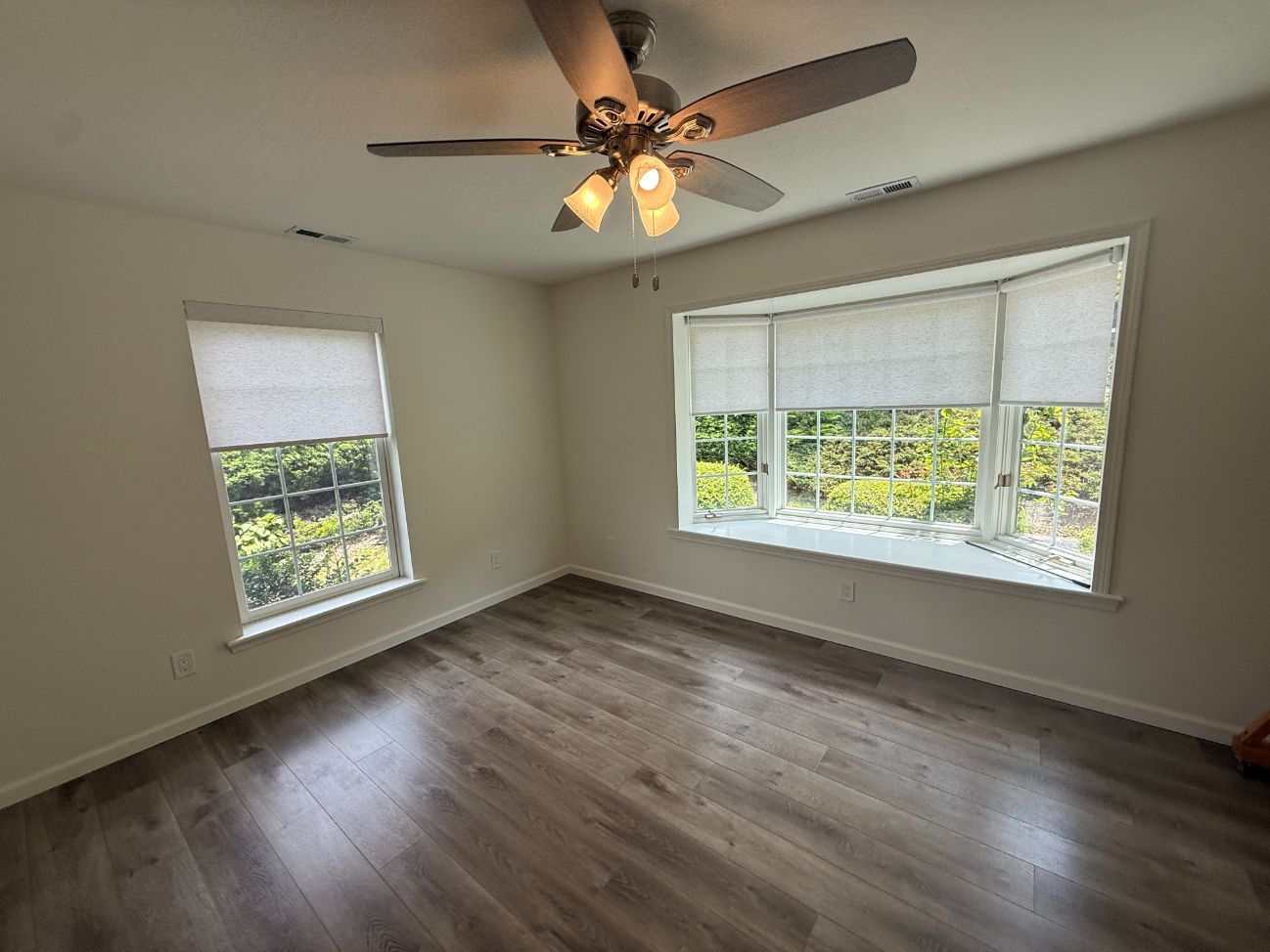 Empty room with wood floors, large windows with roller shades, and a ceiling fan with lights.