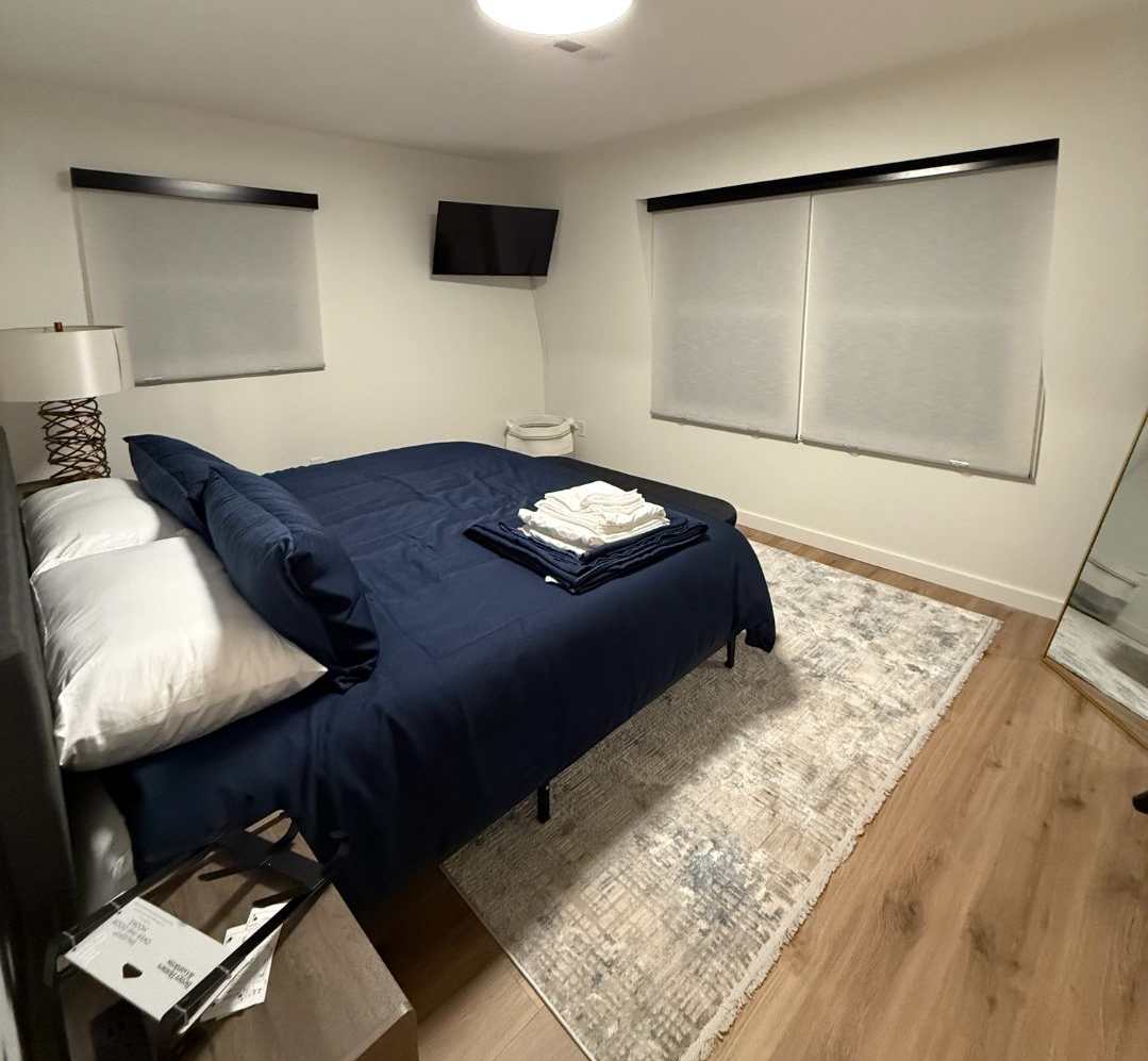 A tidy bedroom with a navy bedspread, white blinds, wall-mounted TV, and light wood floor.