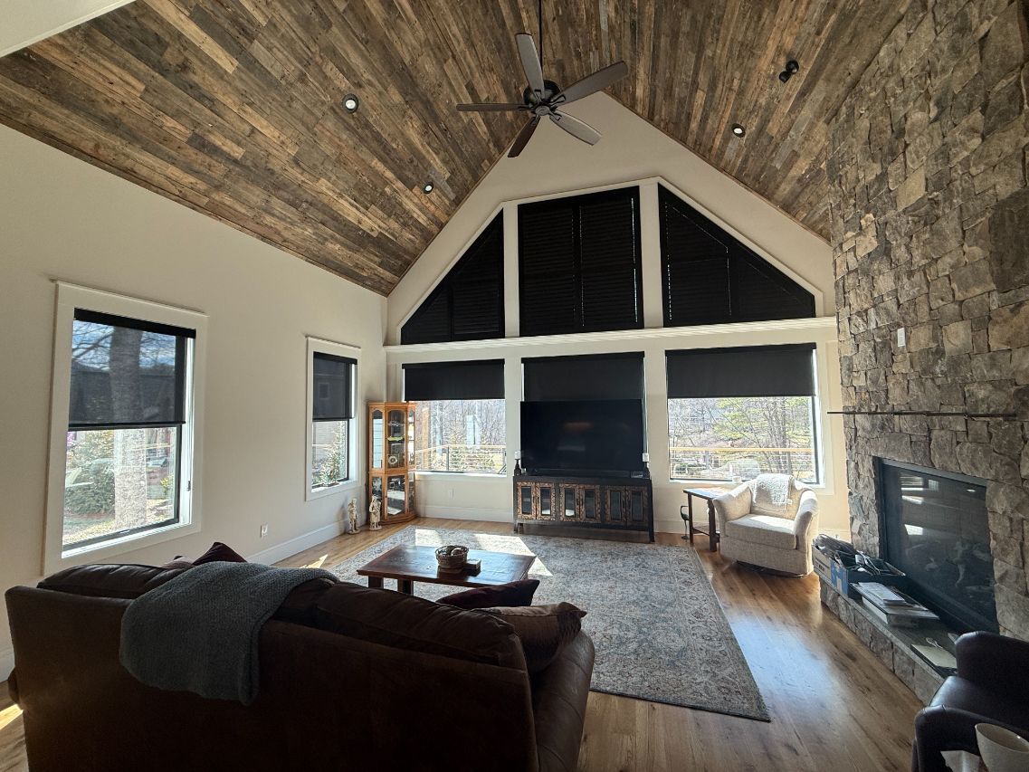 Spacious rustic living room with vaulted wood ceiling, large windows with plantation shutters, fireplace, and dark sectional sofa.