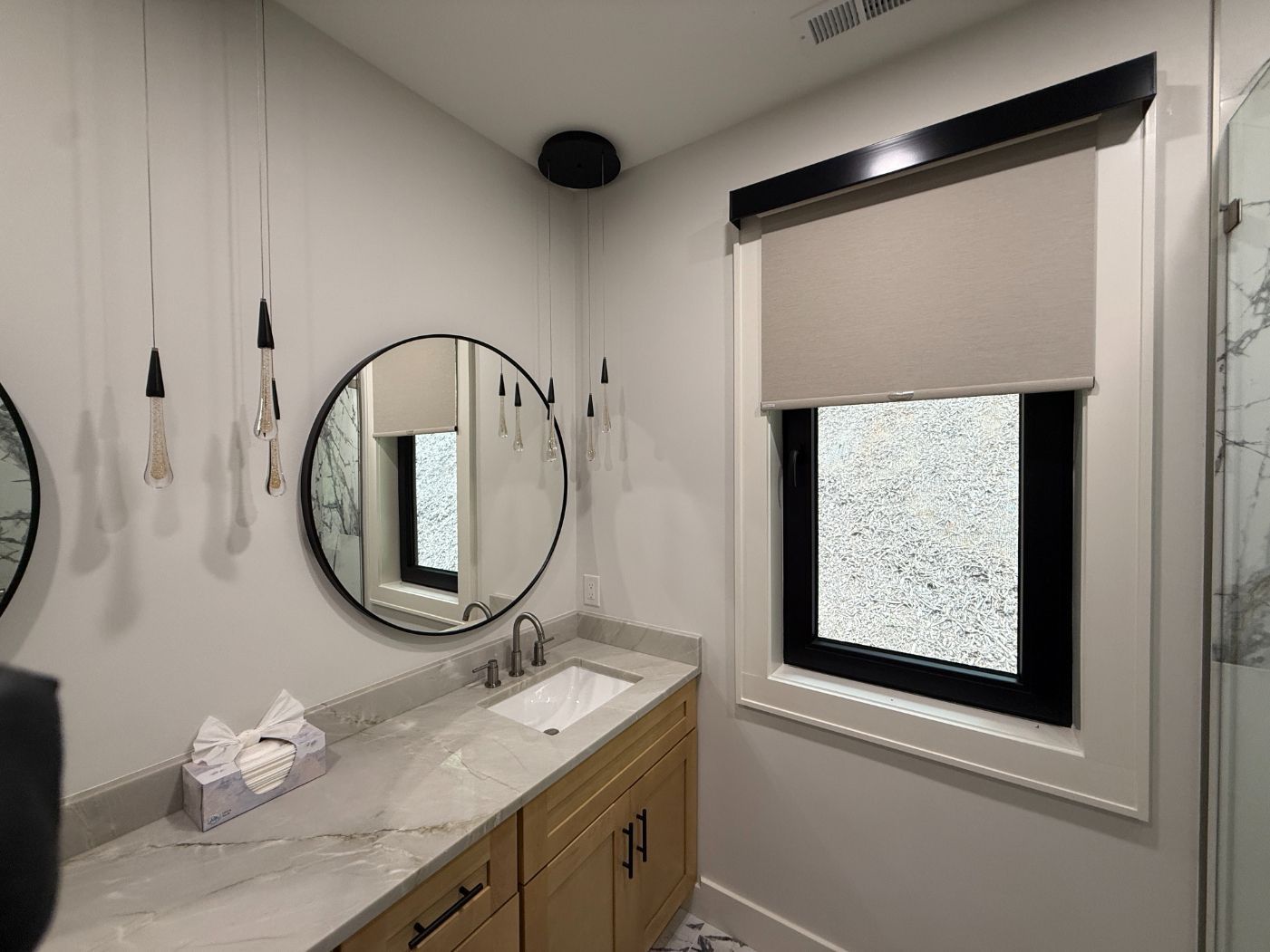 Bathroom vanity with round mirror, marble counter, and window with beige blind.