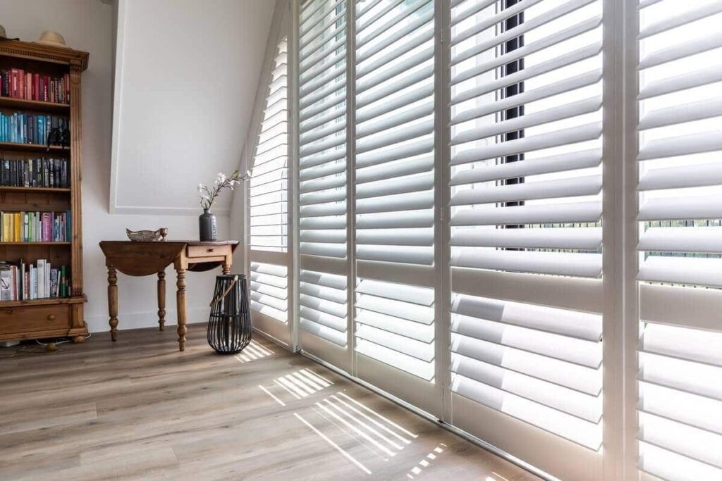 Bright room with white window blinds, wooden floor, and a small desk beside a bookshelf