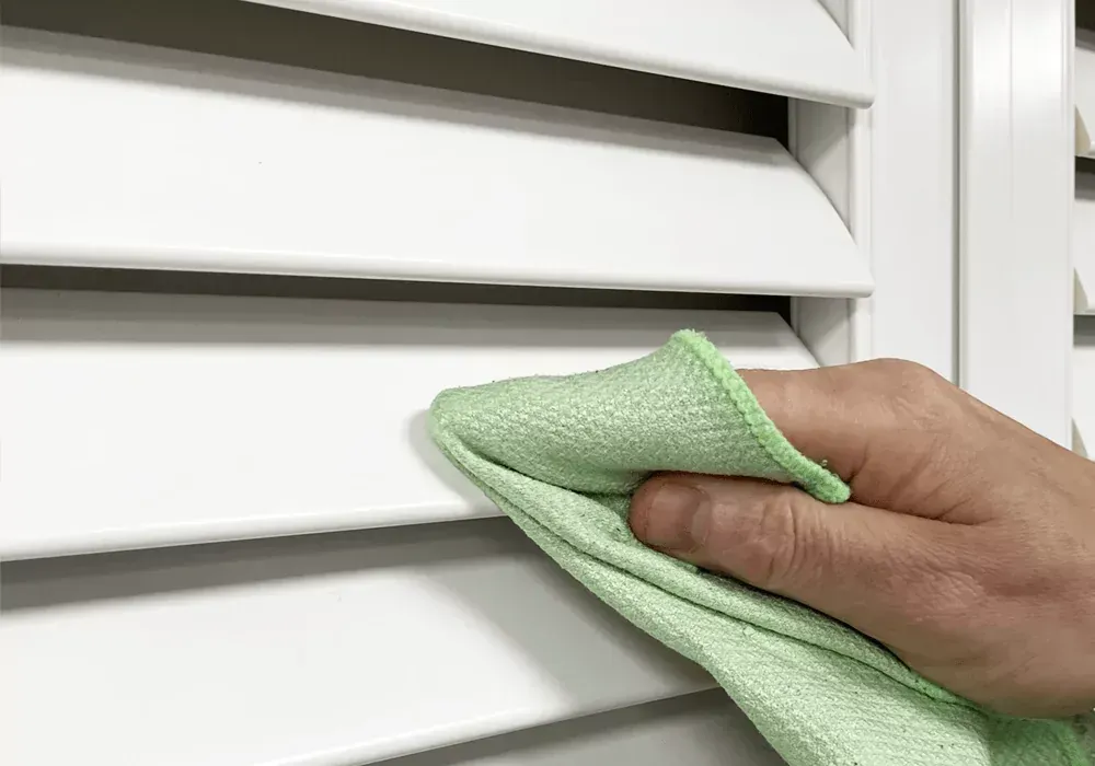 Hand wiping white window blinds with a green cloth