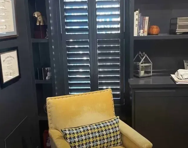 Office with dark shelves, closed shutters, and a yellow armchair with a patterned pillow.