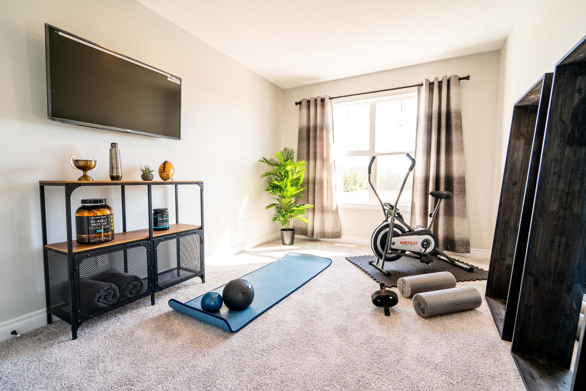 Home workout room with TV, exercise bike, yoga mat, dumbbells, and large window lights.