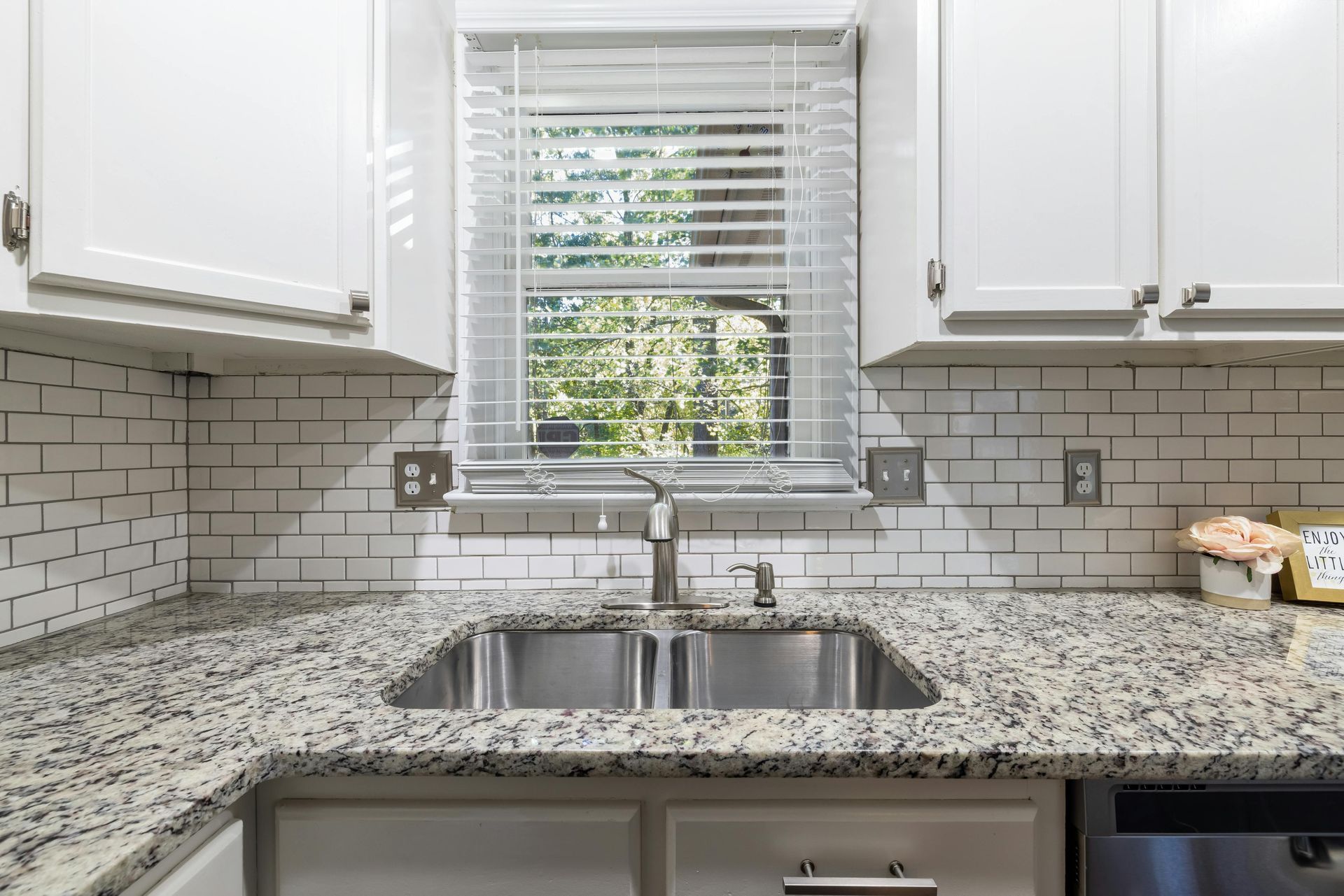 Bright kitchen with white cabinets, speckled granite counters, subway tile backsplash, and a double sink by a window