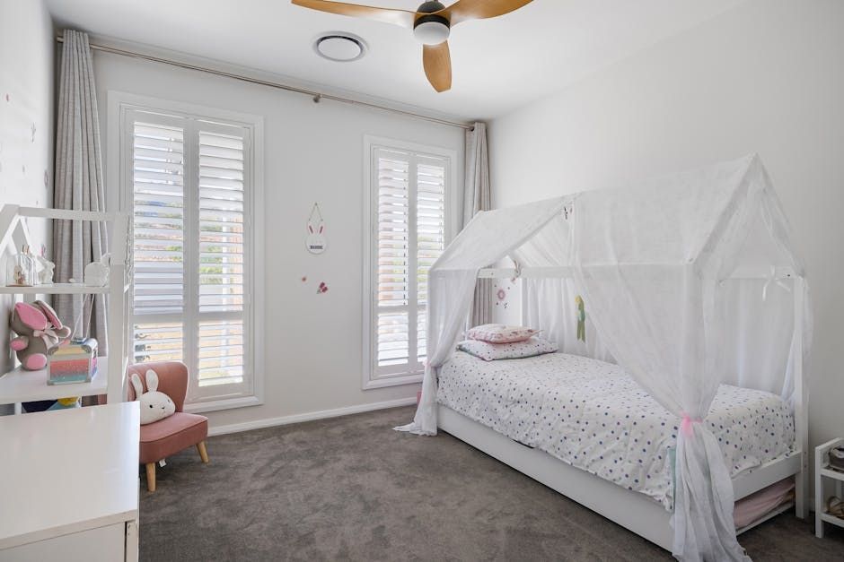 Bright child’s bedroom with a white canopy bed, pink chair, and large shuttered windows.