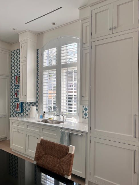 Bright white kitchen with tall cabinets, a window over the sink, and a woven chair at the counter.