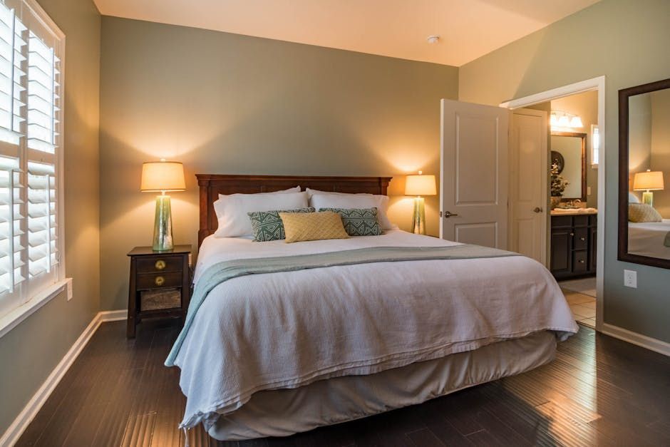 Bedroom with a neatly made bed, bedside lamps, and dark wood floors, lit by warm natural light