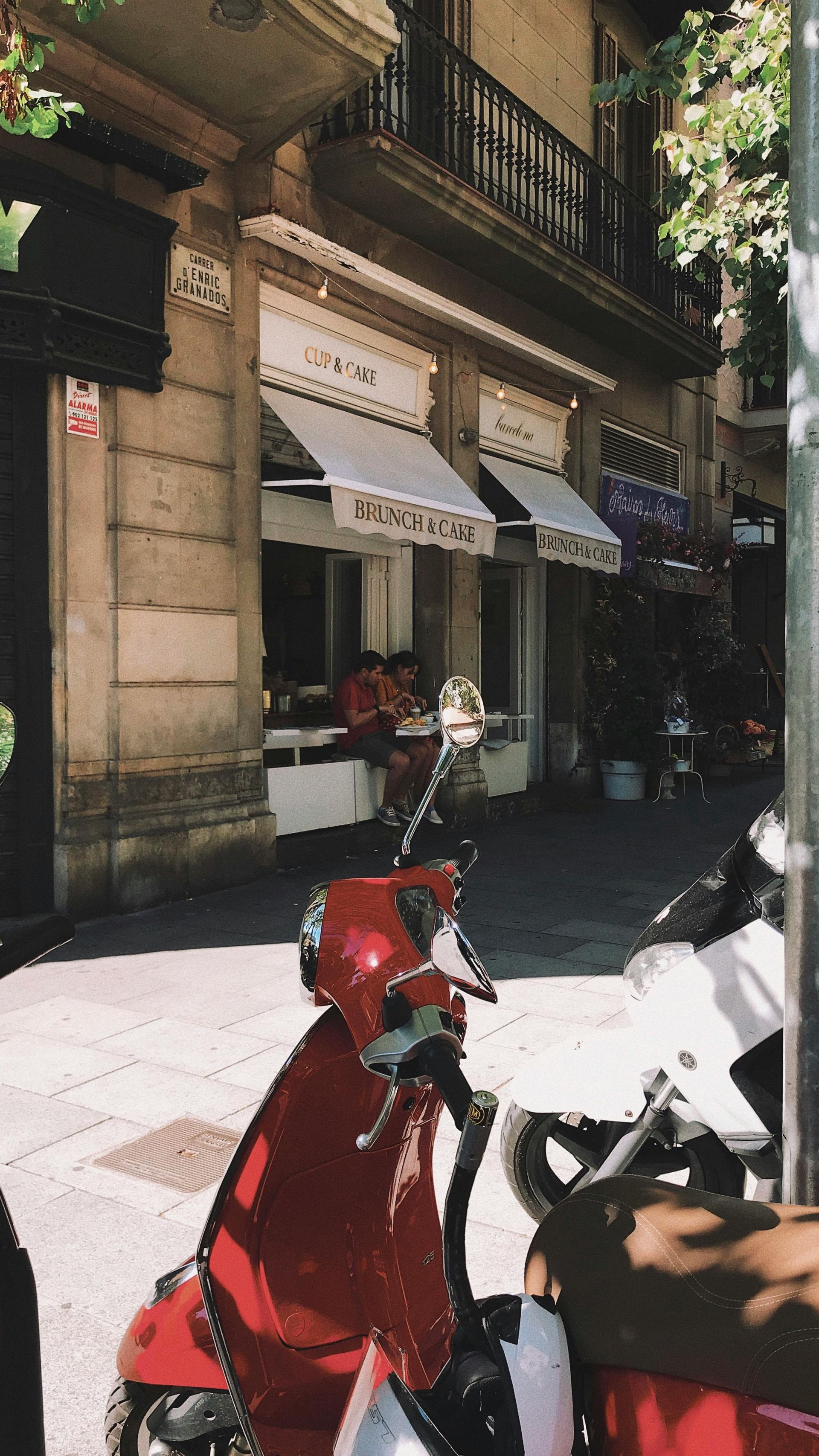Red scooter parked on a sunlit city sidewalk beside a storefront and building facade.