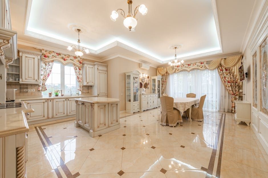 Elegant cream-colored kitchen and dining area with marble floors, island, chandelier lights, and sheer curtains.