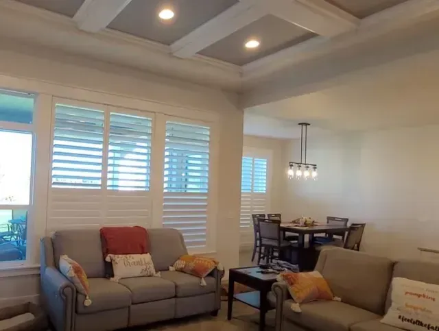 Bright living room with two sofas, dining table, white shutters, and a coffered ceiling with recessed lights