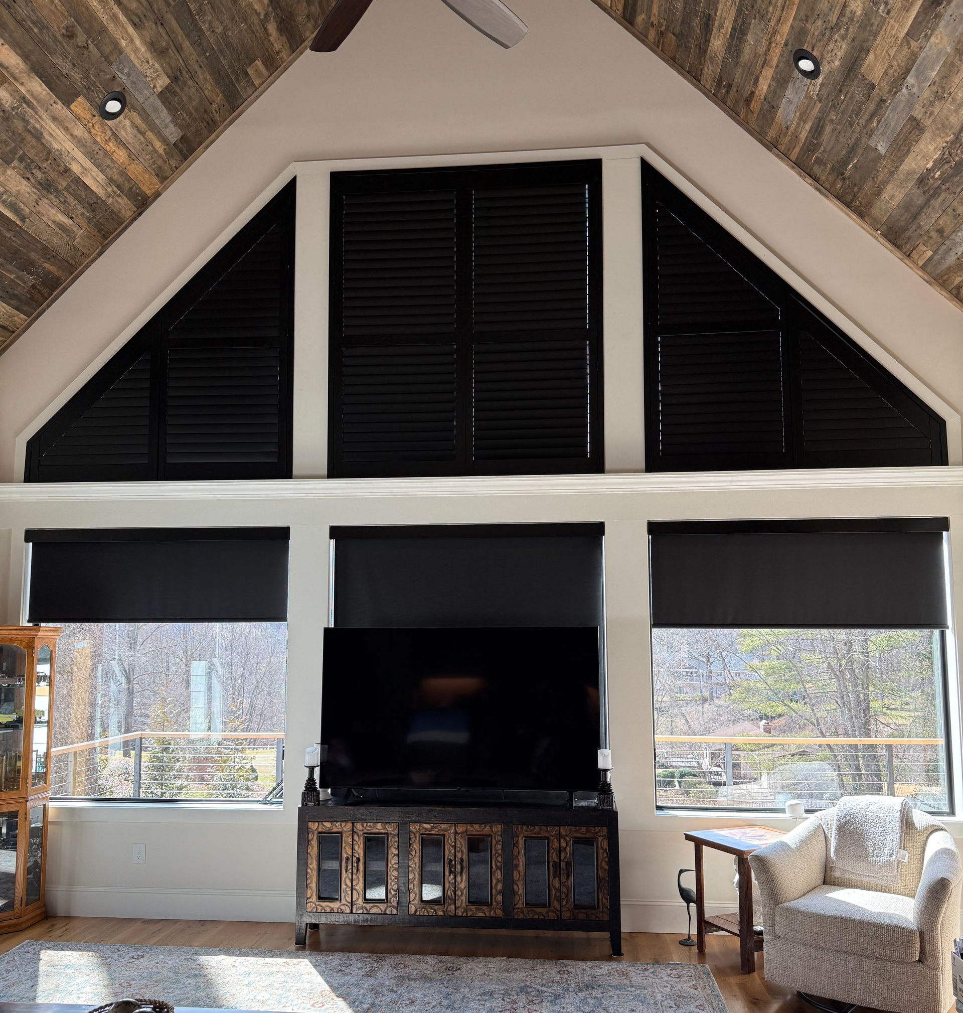 Modern living room with vaulted wood ceiling, black shutters, TV cabinet, and armchair by large windows