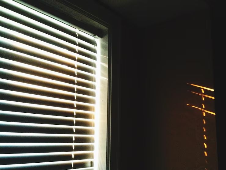 Sunlight streaming through white blinds into a dark room, casting warm reflections on the wall