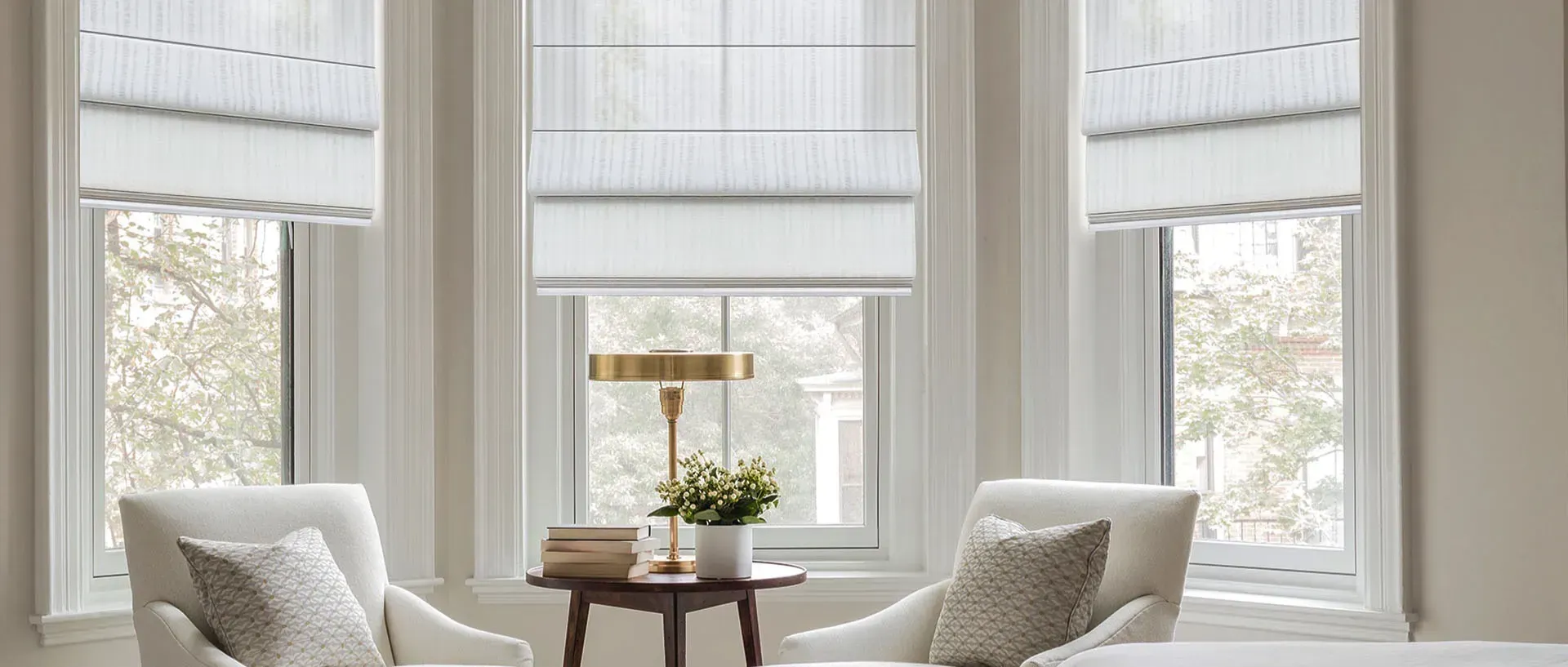 Bright living room with white bay windows, sheer curtains, and two white chairs flanking a small round table.