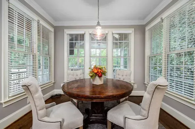 Bright breakfast nook with round wooden table, white chairs, and large windows with blinds