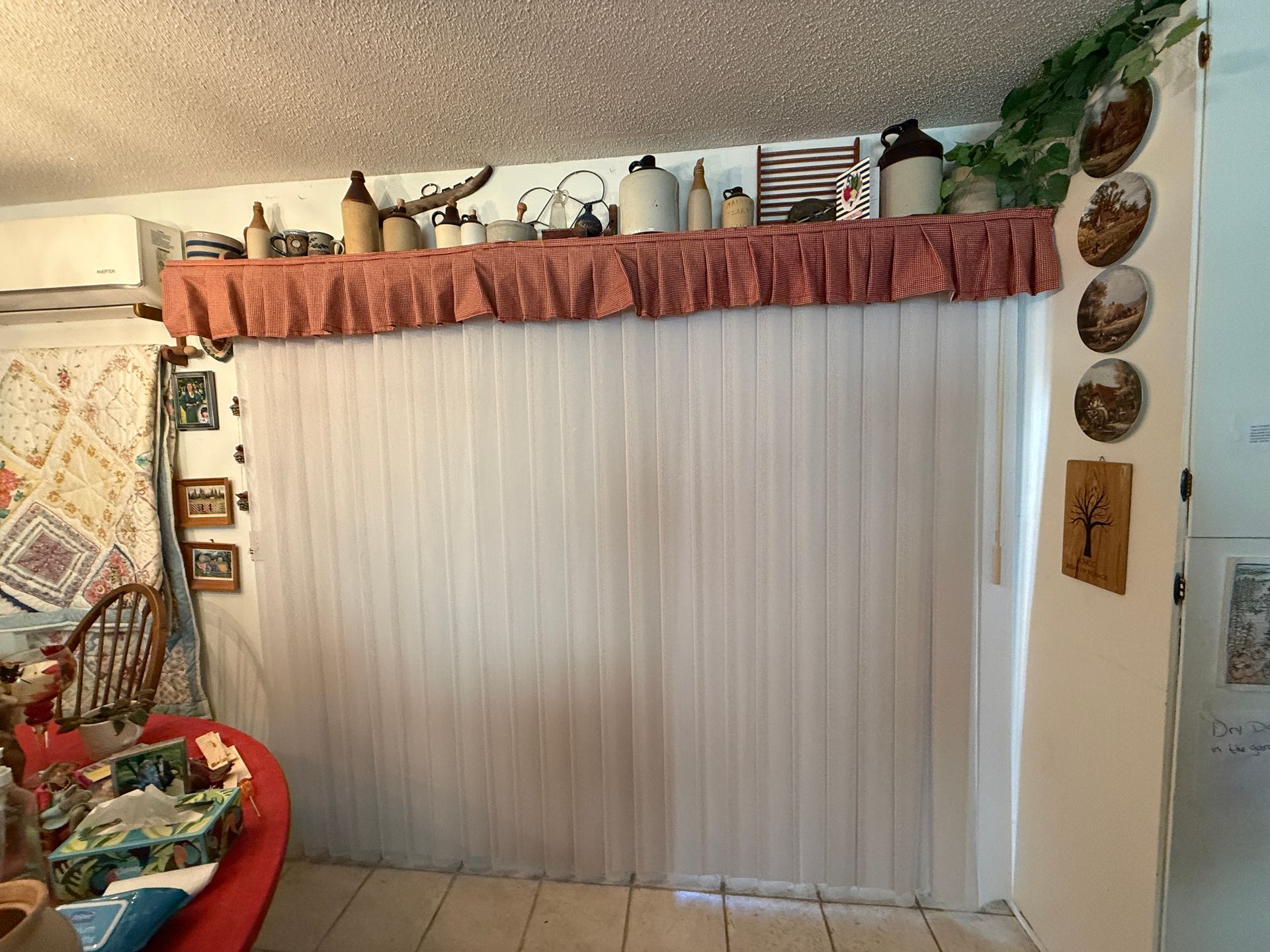 Living room window with white vertical blinds, red valance, and decorative wall hangings above.
