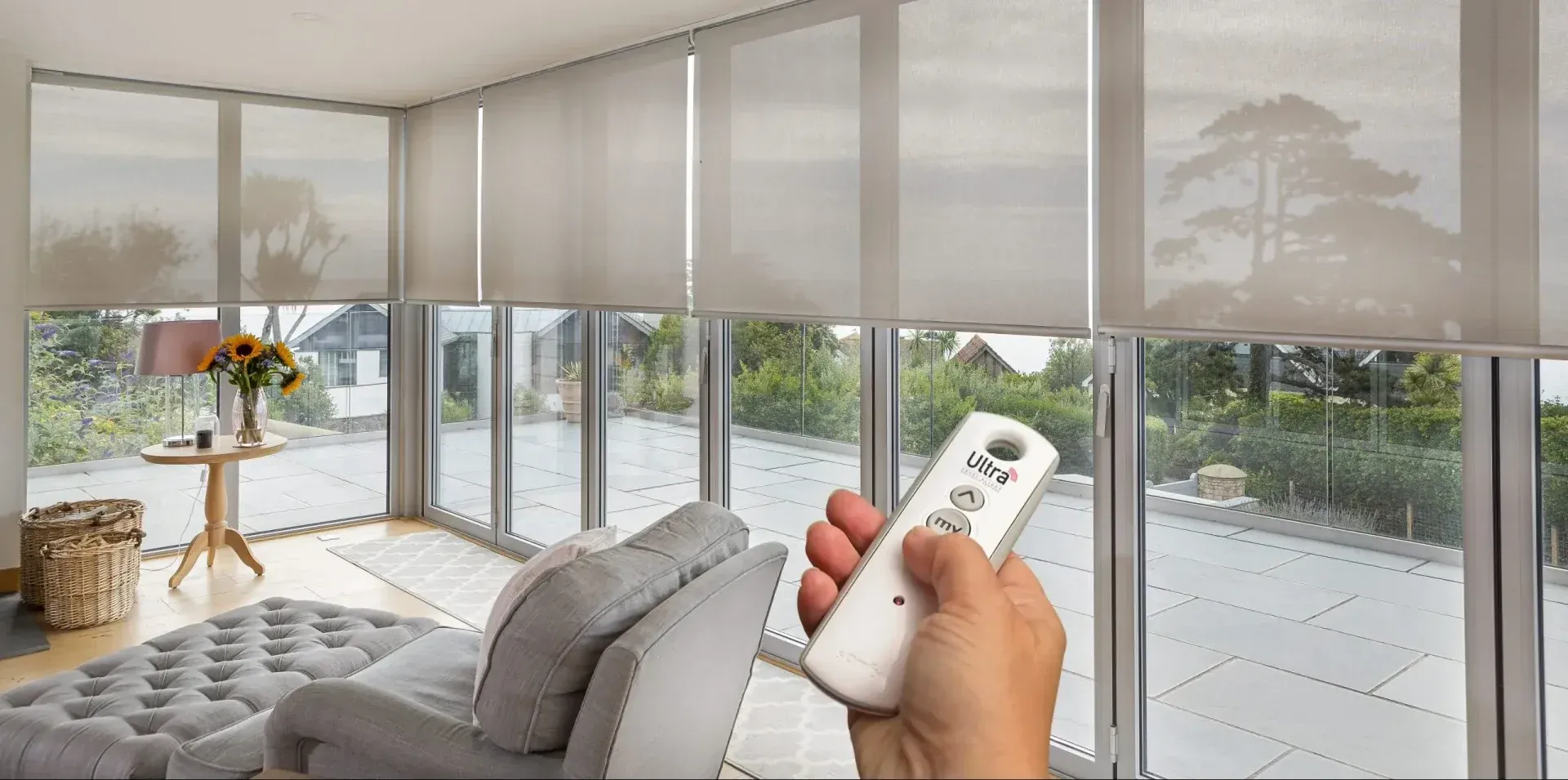 Hand holding a remote toward blinds in a bright living room with large windows and a patio view