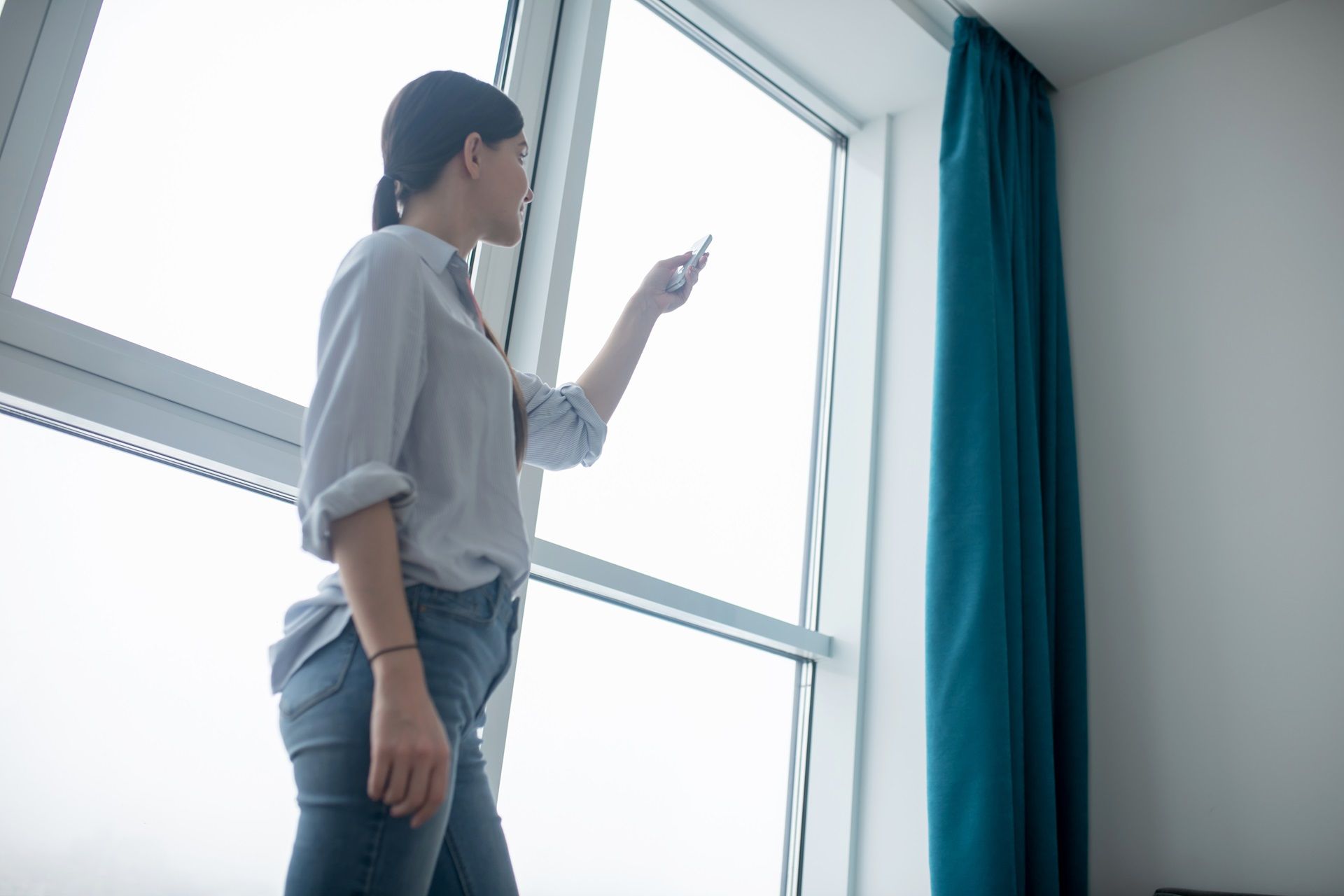 Person standing by a large window, pointing at the glass in a bright room with blue curtains.