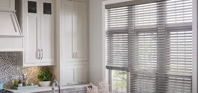 Bright white kitchen with cabinets, sink, and a window covered by gray blinds.