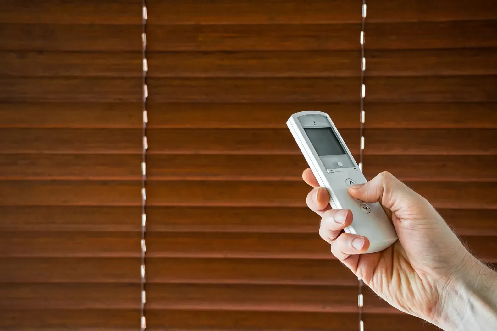 Hand holding a white remote control in front of brown window blinds
