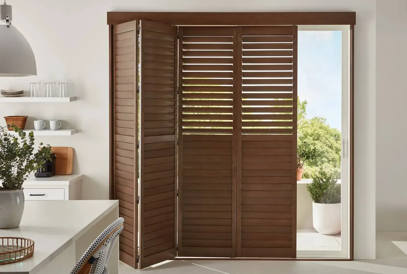 Wooden louvered folding doors beside a bright doorway open to a sunny balcony with greenery.