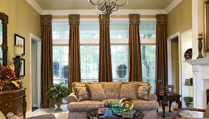 Elegant living room with tall windows, patterned sofa, chandelier, plants, and a white fireplace