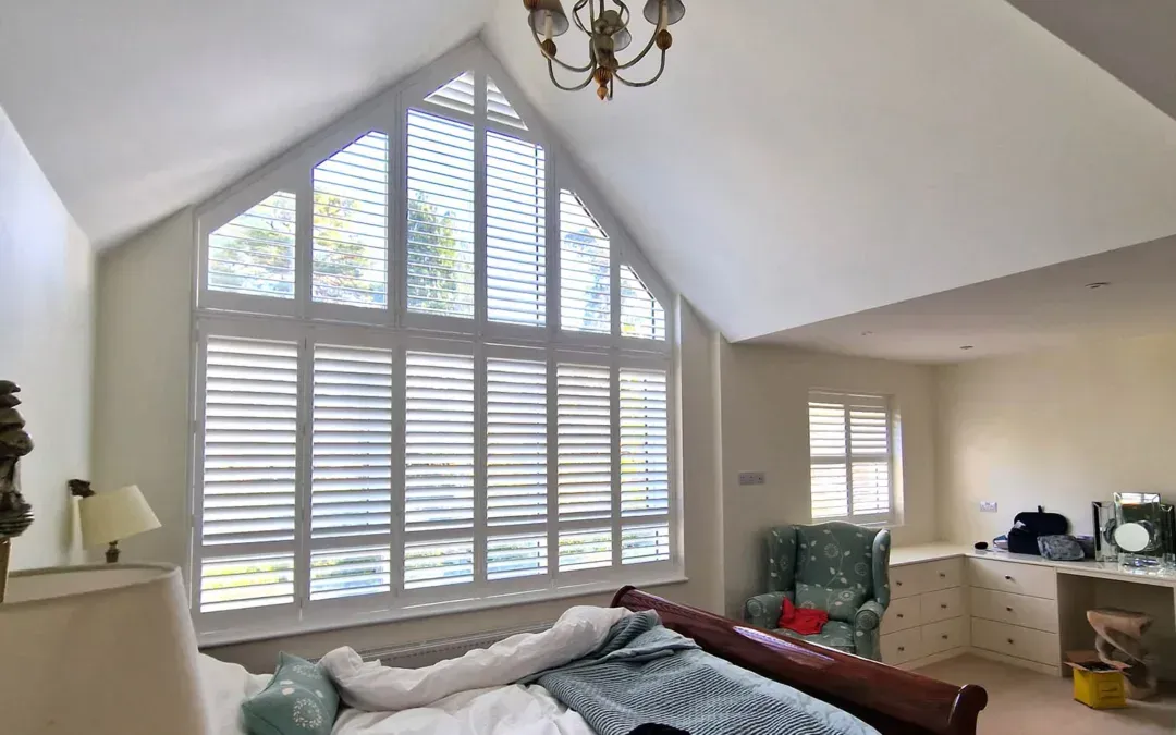 Bright bedroom with vaulted ceiling, large arched window with blinds, dresser, and bed in foreground