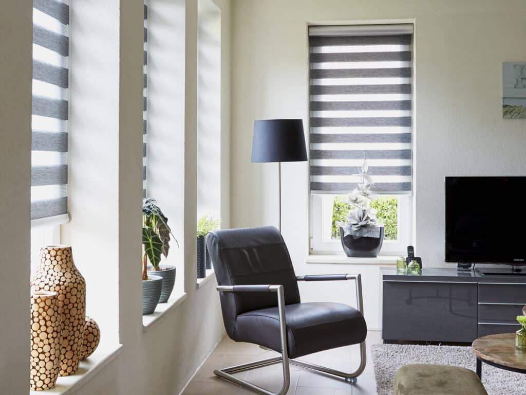 Modern living room with gray chair, floor lamp, striped blinds, TV, and woven decor accents