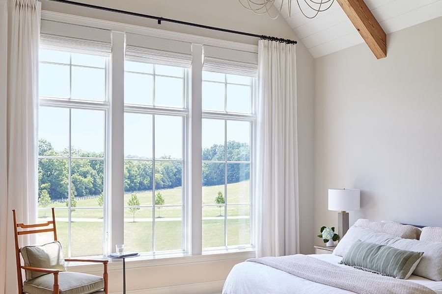 Bright bedroom with large windows, white curtains, a bed, and a chair overlooking a green landscape