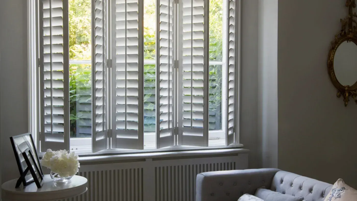 Bright living room with white shutters, a radiator, and a tufted sofa by a round side table and mirror.