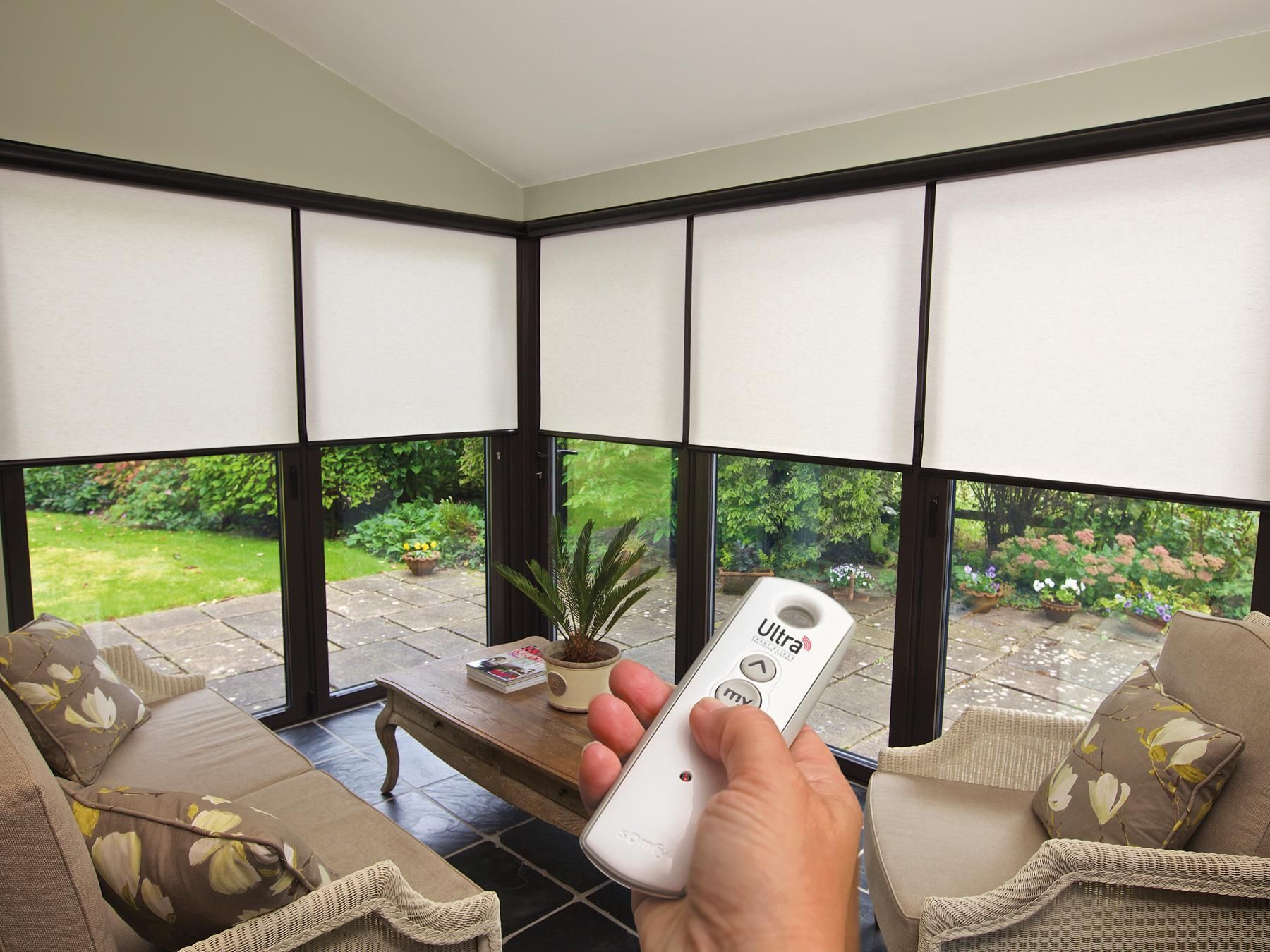 Hand holding a remote in a screened porch with beige chairs and garden view through windows