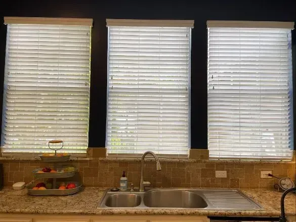 Kitchen sink beneath three windows with white blinds and a fruit basket on the counter